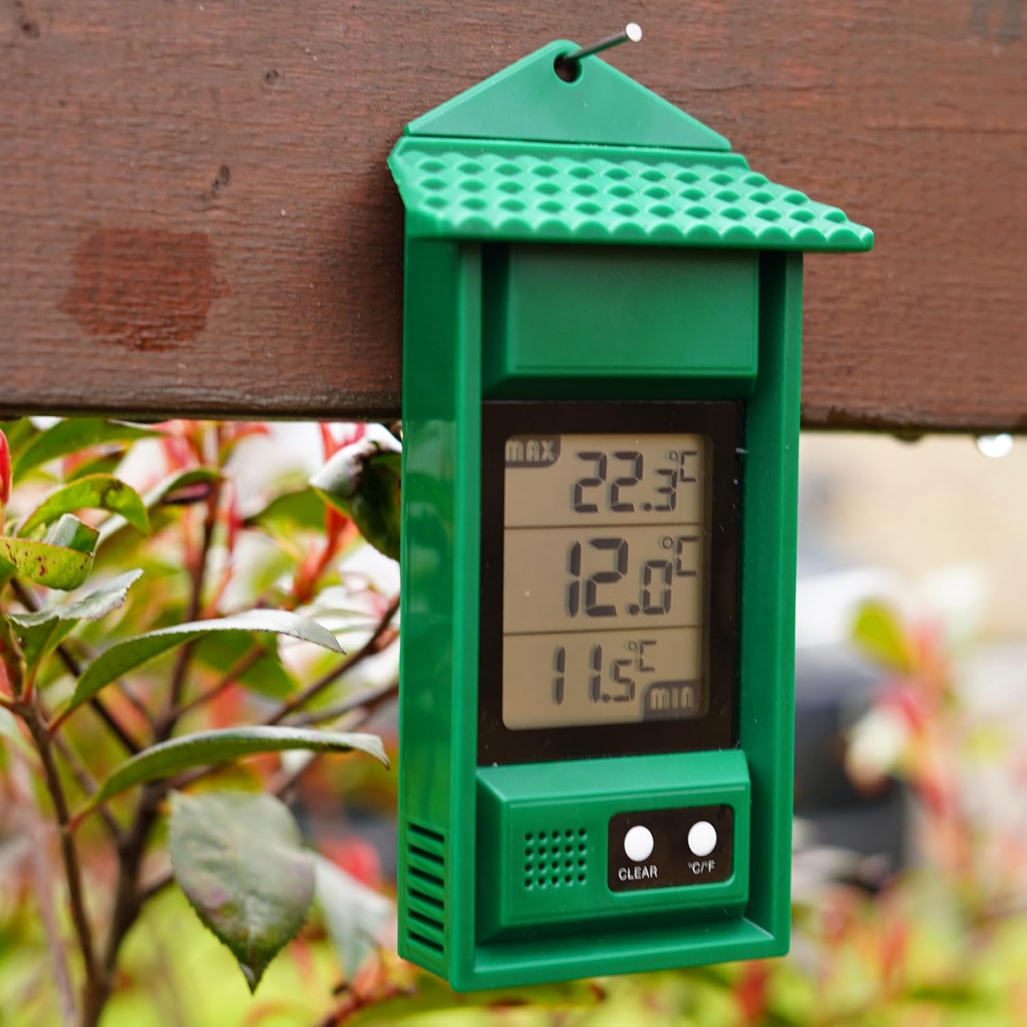The max min thermometer hung from a single nail on a garden fence rail beside red-tipped foliage, showing max 22.3C and min 11.5C