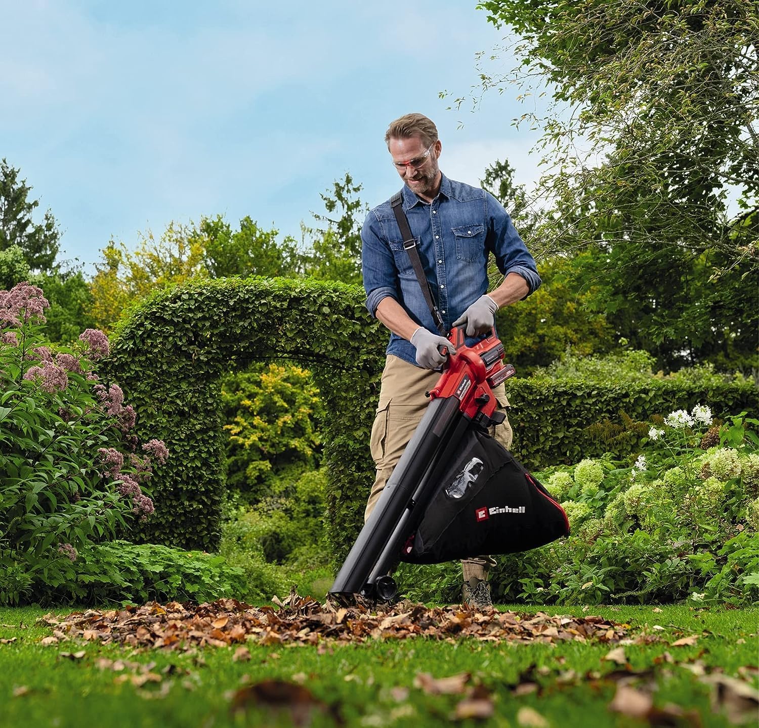 Gardener in clear safety glasses and work gloves vacuuming a pile of dry brown leaves off a mown lawn with the Einhell VENTURRO, the carrying strap over one shoulder and the collection sack at his hip