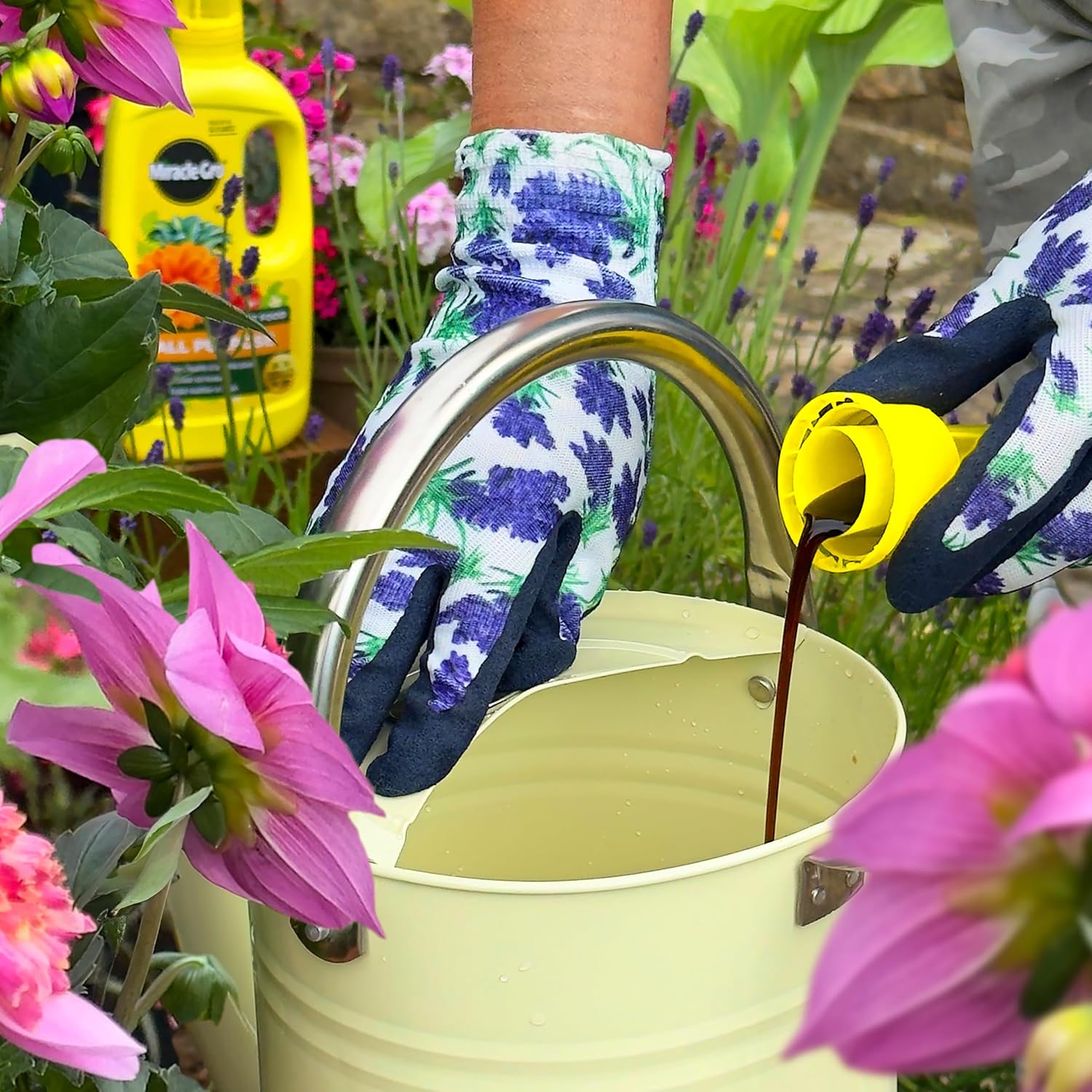 Gloved hands in floral gardening gloves pouring concentrated plant food from the measuring cap into a cream metal watering can