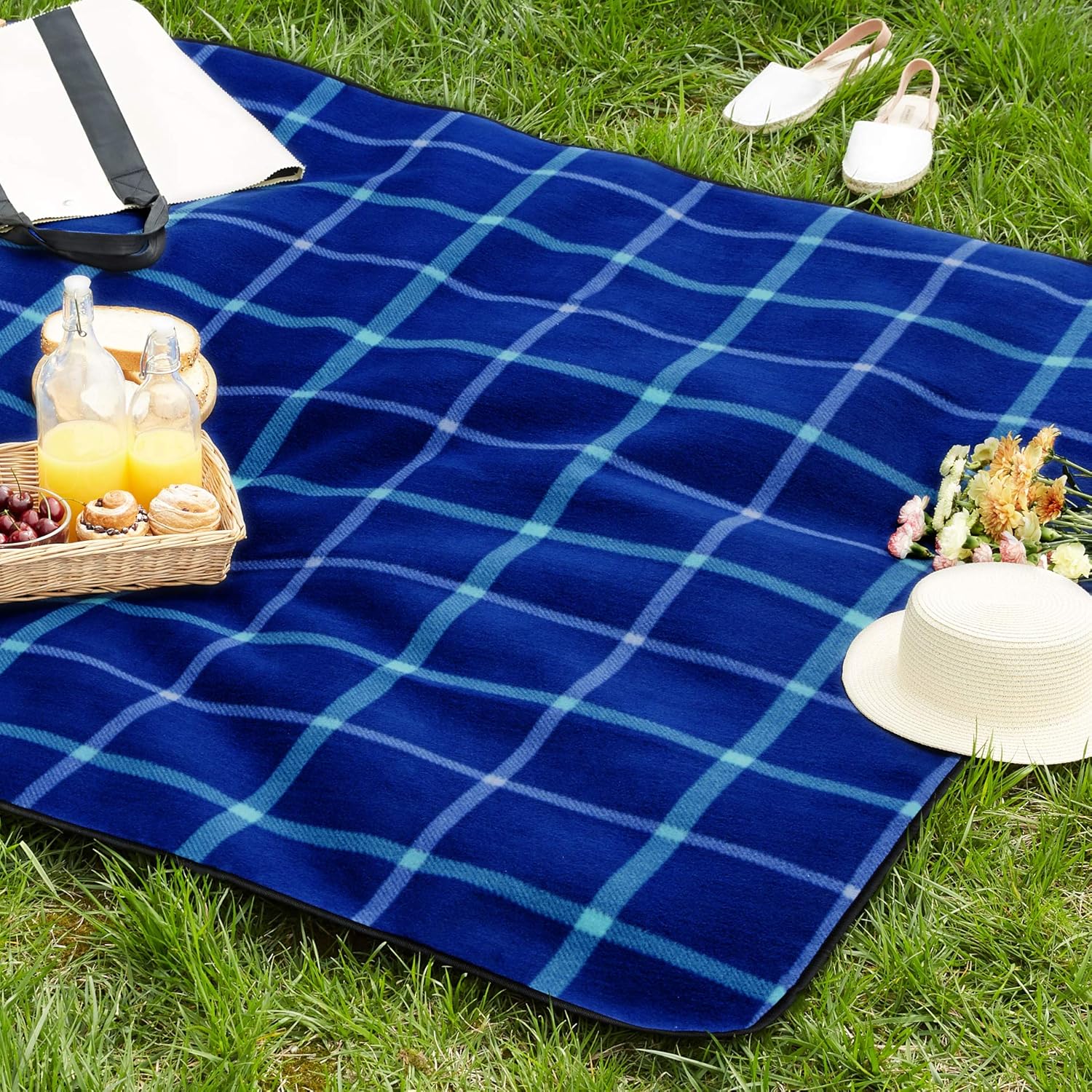 Amazon Basics blue check picnic blanket spread on grass with a picnic basket, drinks and sun hat on top