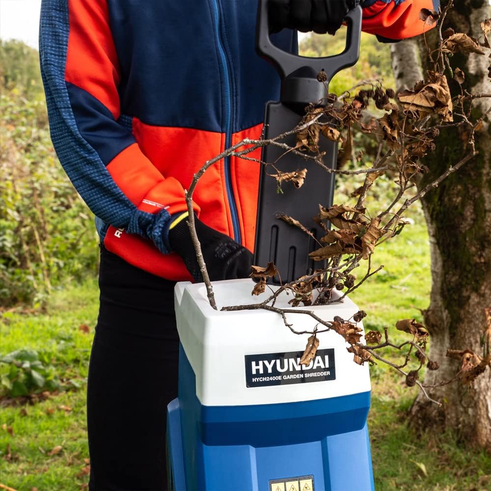 Gloved hands feeding a dry brown-leaved branch into the hopper of the Hyundai shredder with the flat push stick resting in the mouth